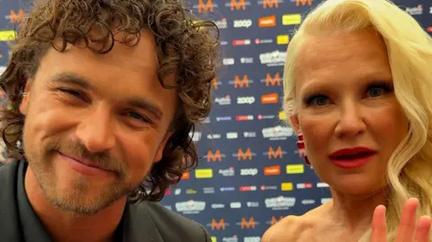 A smiling man with curly brown hair and a woman with long blonde hair look at the camera