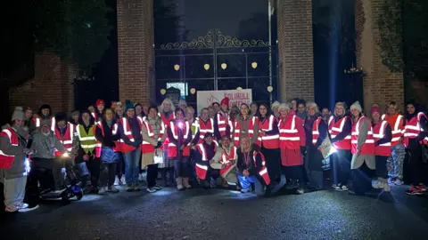 BBC A group of around 100 women smiling into the camera wearing pink high-vis jackets and all looking into the camera. They stand in front of red brick pillars which are either side of gates.
