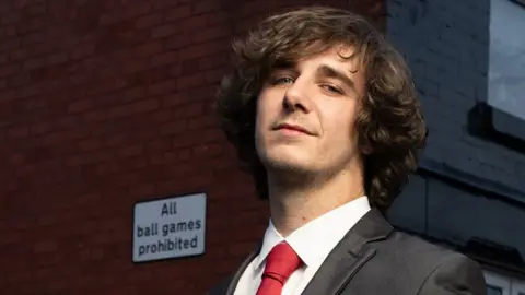 PA Media Brad Kella, with mid-length curly brown hair, wears a dark suit with a red tie as he stands in a Liverpool street of terraced houses with a sign on the wall reading "All ball games prohibited"