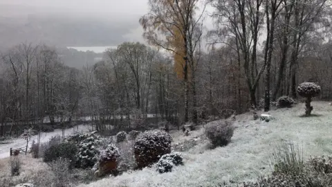 Snow-covered garden and trees on a hillside, with a misty lake and forest in the background under an overcast sky.