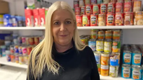 BBC/Heidi Tomlinson A blonde woman standing in front of shelves of tinned food