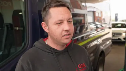 Joshua Hough stands in front of a blue van. He has short brown hair and is wearing a black hoody with red writing. 