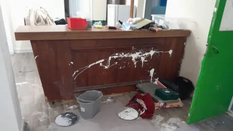 A vandalised piece of furniture in a clubhouse. White paint has been thrown across it and items are cluttered on top of the unit and on the floor.
