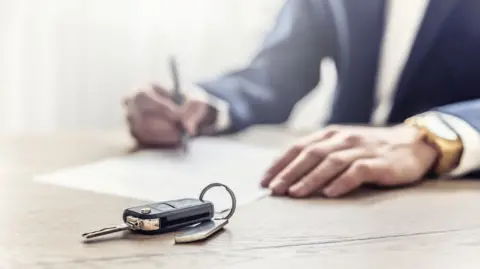 Getty Images Man in a suit writing on a piece of paper with car keys in focus in front of him.