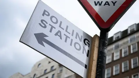 A white polling station sign with a black arrow on it attached to a sign post. Buildings can be seen towering above the sign from a distance away.