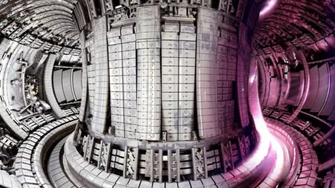 UKAEA JET reactor at Culham before it was decommissioned. It is like being inside a big hollow donut made of incredibly shiny metals. A pink wave of energy is passing through it from the right hand side.