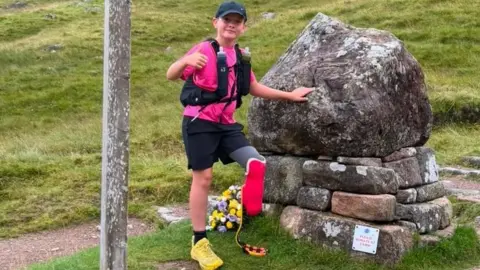 Daniel Thomas A 9 year-old boy with his left foot amputated. He is climbing a mountain and is photographed during his climb. He is wearing one bright yellow trainer, black shorts, a bright pink top and a vest, carrying two drinks bottles. He is holding his thumb up, with his other hand placed on a pile of rocks, smiling and looking at the camera.