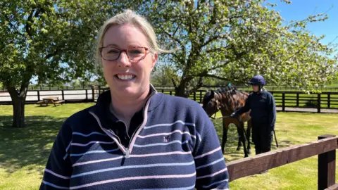 Gemma Lamble smiling at the camera. She is wearing a striped jersey and glasses. Behind her is a fenced paddock with trees, grass and a young woman holding a horse by its reigns. She is wearing a riding helmet. 
