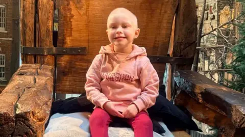 A little girl wearing a pink jumper and leggings smiling and sat on a giant wooden chair