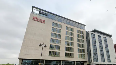 Google The exterior of a nine-storey building with a lamp post in front of it, and a sign that reads "Hilton Garden Inn"
