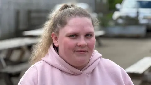 A young woman smiling at the camera. She has her hair in a ponytail and is wearing a pink hoodie. She's sitting on a picnic bench area at Forge Mill Farm in Sandwell. 