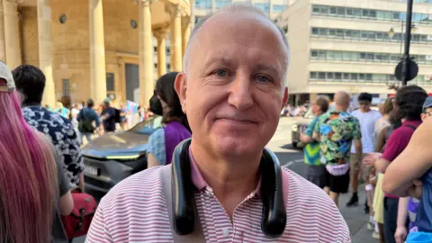 Simon, a middle-aged man with grey hair, smiles as he is pictured wearing a striped pink and white polo shirt, and a portable neck fan, as people gather ahead of the march