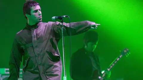 Oasis lead singer Liam Gallagher in a parka jacket pointing at the crowd in front of a microphone with his brother and bandmate Noel behind him, face down playing a guitar on stage in 2009 with green lights lighting the stage 