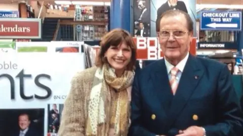 Anika Pavel Anika Pavel wearing a scarf and cream coat smiling at the camera. She is standing next to Roger Moore who is wearing a navy suit