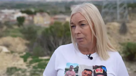 Sharon George. She has straight blonde shoulder length hair, wearing a white t-shirt with images of John on it.