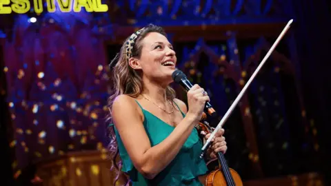 EIF Close-up of a musician on stage at the Edinburgh International Festival holding a violin and microphone, with a bright yellow “Edinburgh International Festival” sign glowing behind and ornate architectural details lit in blue in the background.