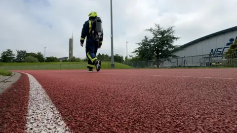 Nick Hays is walking on a red tarmacked race track in his full fire fighting kit, dark blue suit, with yellow fluorescent strips, cylinder on his back and yellow helmet.