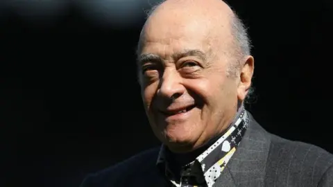 Getty Images A close-up of Mohamed Al Fayed's head and shoulders. He smiles at the camera and wears a dark gray suit with stripes and a patterned shirt.