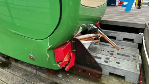 The front of a green train has been crushed in by the buffer, which is a piece of red and white painted wood that has splintered.