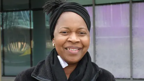 Progress Mushonga is smiling at the camera. She has brown eyes and and is wearing gold small hooped earrings and a black coat with a black scarf. 