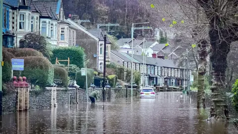 Emma Peterson Berw Road in Pontypridd with  the road underwater, and a car up to its wheel well. A person can also be seen standing almost up to their knees in water. 