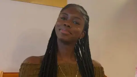 Naomi Oyebode wearing green top and gold necklace sitting on an orange sofa