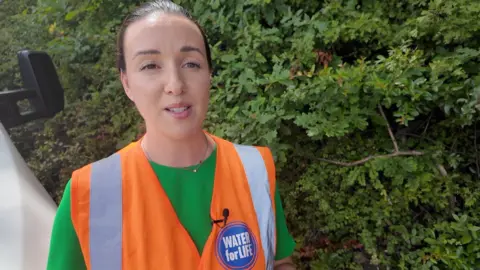 A woman wearing a green top and a fluorescent orange tabard stands in front of a hedge
