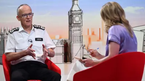 Mark Rowley sits infront of Laura Kuenssberg, who has her back turned to the camera shot. Rowley is wearing his white policing shirt with black patches on the shoulders. Kuenssberg is wearing a light purple dress and they are both sat on red chairs