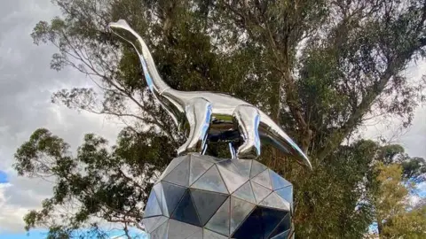 A steel dinosaur sculpture stands on top of a geometric ball in New Zealand.