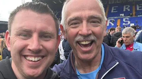 Ash Lambell Ash Lambell and Kevin Odams among other fans on the pitch at a game. They look jubilant, smiling in this selfie. A large stand with blue seats is in the background.