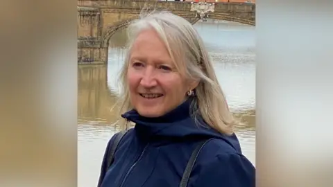 A woman with blonde hair is wearing a navy blue coat and standing in front of a river and a bridge.
