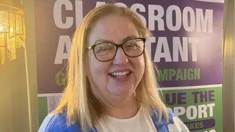 Una mujer sonriente de cabello largo y rubio está de pie frente a un cartel publicitario. Lleva gafas de montura oscura y una camiseta azul y blanca.
