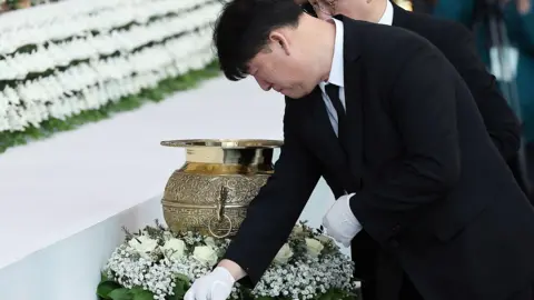 NEWS1 A man in a black suit and white gloves lays a flower down on a table next to a wreath and a golden urn