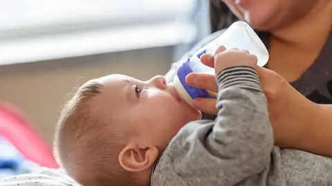 A close up image of a woman feeding a young baby with a plastic bottle.  The baby is in the foreground and wearing a grey sleepsuit.