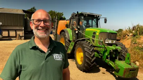 A man with a beard and glasses smiles into camera. He has a green top on. Behind him is a green tractor.