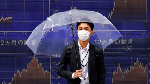 Getty Images Main holding umbrella in front of stock market charts in Tokyo.