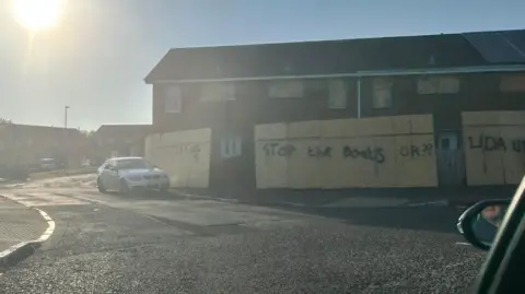 Hoarding outside a set of terrace houses sprayed with graffiti reading: STOP THE BOATS'. A car is parked to the left. 