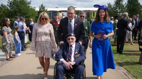 Reuters An old man in a blue suit, wearing military medals, sits in a wheelchair as he is pushed by Prime Minister Kier Starmer. Two women walk alongside Starmer.