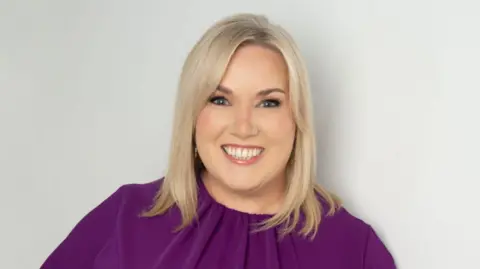 Education Authority A woman with shoulder-length blond hair smiles into the camera. She is wearing a purple dress and is standing in front of a grey backdrop. 