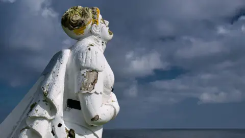 Getty Images The figurehead is white with yellow hair. There a pieces of it that have fallen off exposing wood underneath. The figurehead is next to the sea, which is visible in the distance under a dark blue sky.
