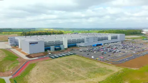 A sprawling grey box building, which is four storeys high, stands next to a car park. The building has an Amazon sign in the corner and is surrounded by fields.