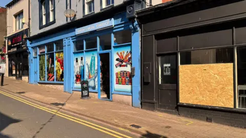 A row of shops on Senhouse Street, Maryport, where the van hit the girls. The shop on the far right has had its window boarded up. It has opening times written on the glass door and a white seat can be seen through the window. The shop looks closed. To the left there is a blue off-licence with an open door. The windows are covered up in photos of crisps, drinks and chocolate bars. There is a takeaway shop on the far right.
