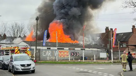 Large flames and plumes of black smoke can be seen coming from a car wash facility behind a metal fence. Emergency service workers and a fire engine are on the scene.