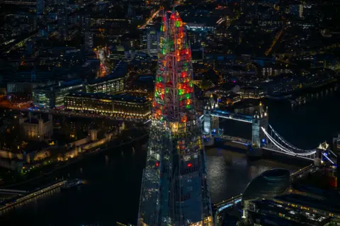 Jason Hawkes Aerial photograph taken at night showing red, orange and green lights at the top of the Shard with the Thames and Tower Bridge lit up behind it