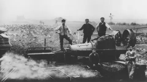 William Blyth An old black-and-white photograph of five workers posing around a large industrial machine in an open excavation site. Three workers stand behind the machinery while two sit or lean in the foreground. Narrow rails run toward the machine, and piles of earth surround the trench. The flat rural landscape stretches into the distance, with a few faint buildings and structures visible through haze in the background. The image has an early-industrial-era appearance.