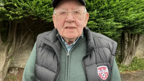An elderly man looks into the camera. He is wearing glasses and a baseball cap with a green and white checked shirt, a light green zip-up cardigan, and dark sleeveless jacket with red-white-and-black Derry City logo on the breast.