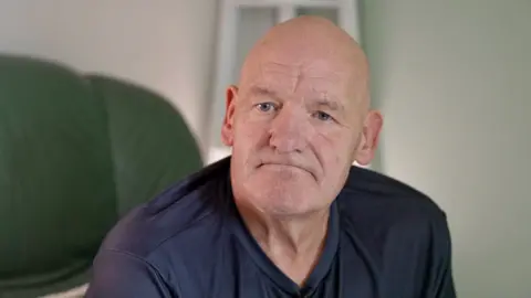 Patrick Moore sitting on a green chair looking at the camera wearing a navy blue top. He has a bald head and is looking glum at the camera.