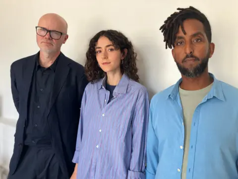 Three people stood in a row, from left - a bald man, wearing black framed glasses, navy suit, and navy shirt, a woman in pinstripe blue shirt, long dark wavy hair and a man with dreads, shaved hair at the side and blue shirt, green T underneath.