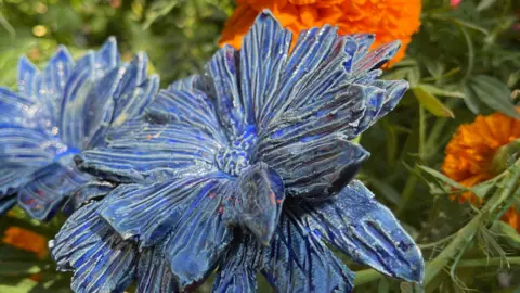 A clay flower in blue - carved petals. Blurred behind is green foliage and real orange flowers.