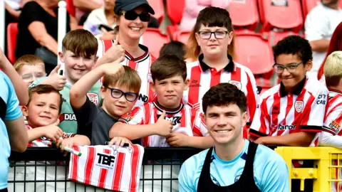 Kevin Morrison A number of young fans stand in front of a row of stadium seats. in front of them is a footballer. The kids are in football kits.